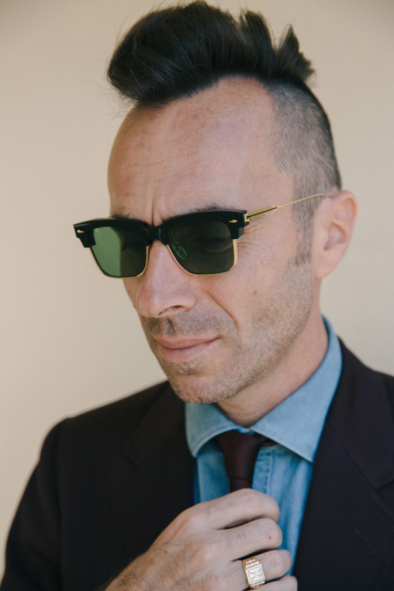Man wearing black and gold-trimmed sunglasses with green lenses, black blazer and blue dress shirt.