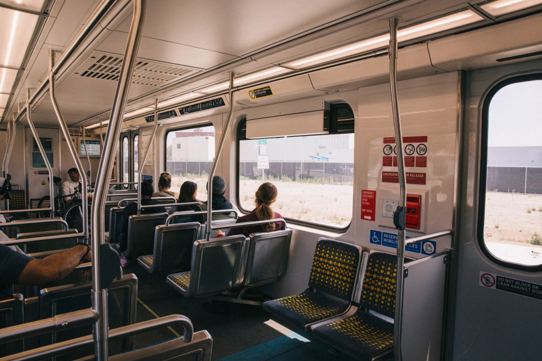 Interior of a light rail subway car with passengers seated and windows showing an industrial area outside.