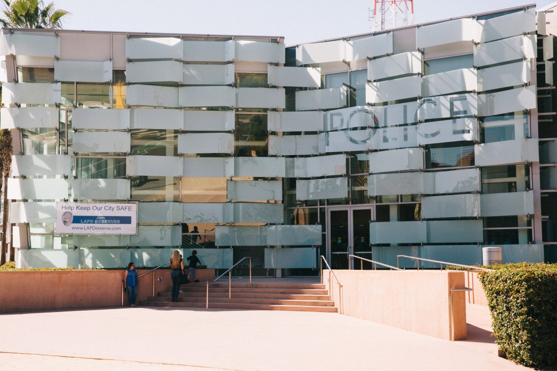 Modern white and glass building facade with horizontal striped pattern, steps leading to entrance, palm trees visible.