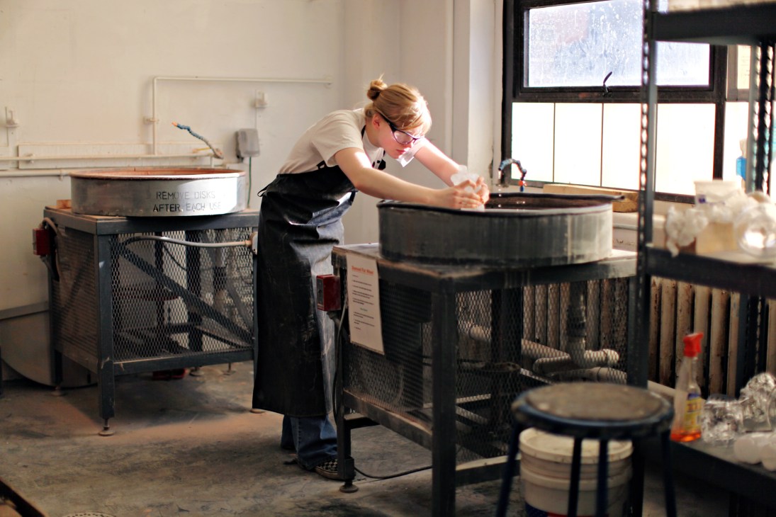 A student wearing glasses and an apron works with molten glass at a furnace in a studio workshop.
