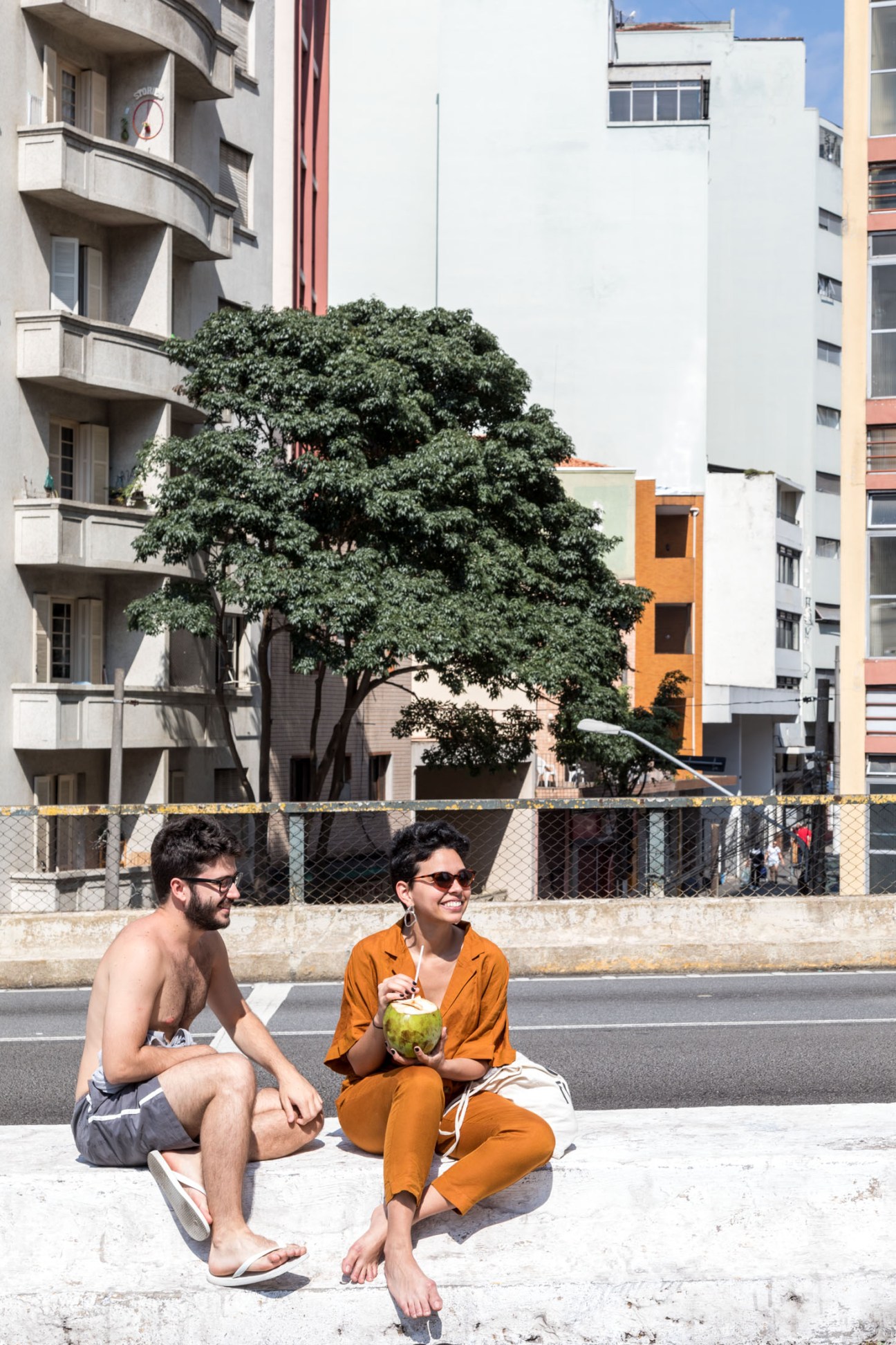 Two people sitting on Minhocão Park's elevated walkway with coconut drinks, surrounded by São Paulo buildings.