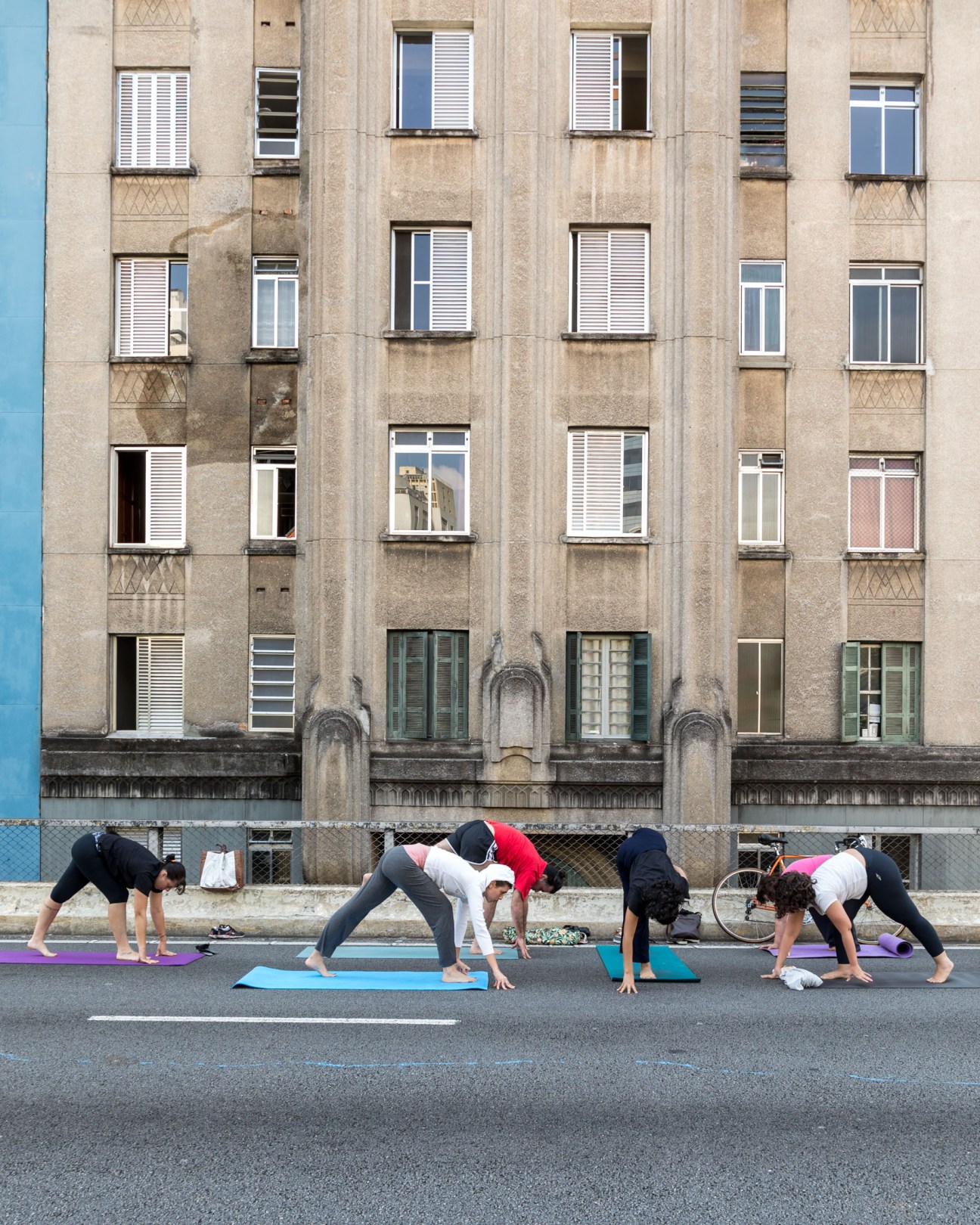 People doing yoga poses on colorful mats on a street in front of a beige apartment building