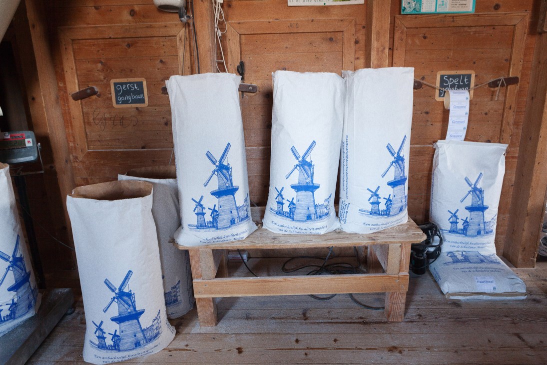 Sacks of barley and flour with blue windmill prints stacked on wooden shelf in rustic wooden interior.