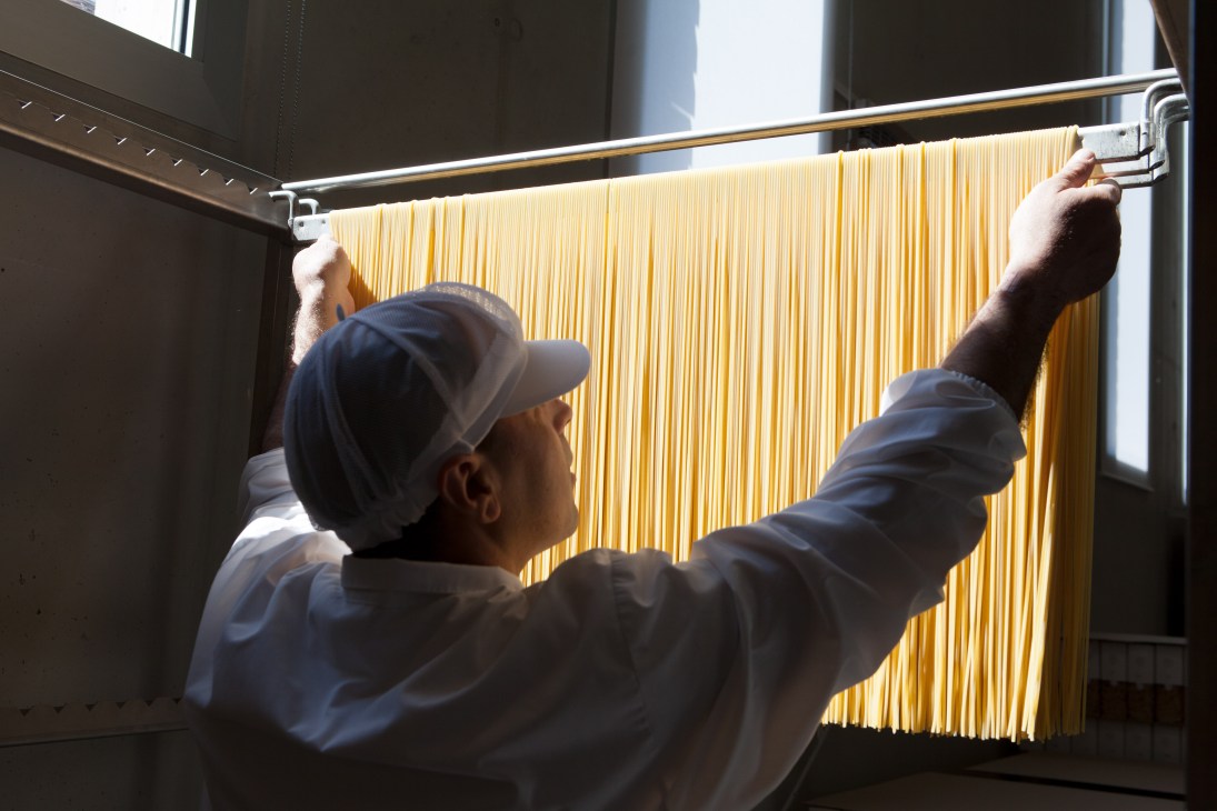Worker hanging long strands of spaghetti on a metal rod for air-drying in a pasta factory.