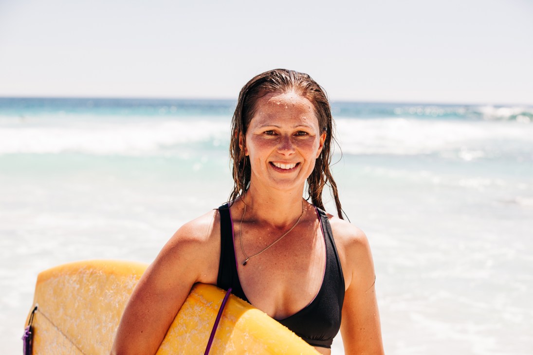 Surfer Sarah Annersley smiling at camera while holding yellow surfboard on beach with ocean waves in background
