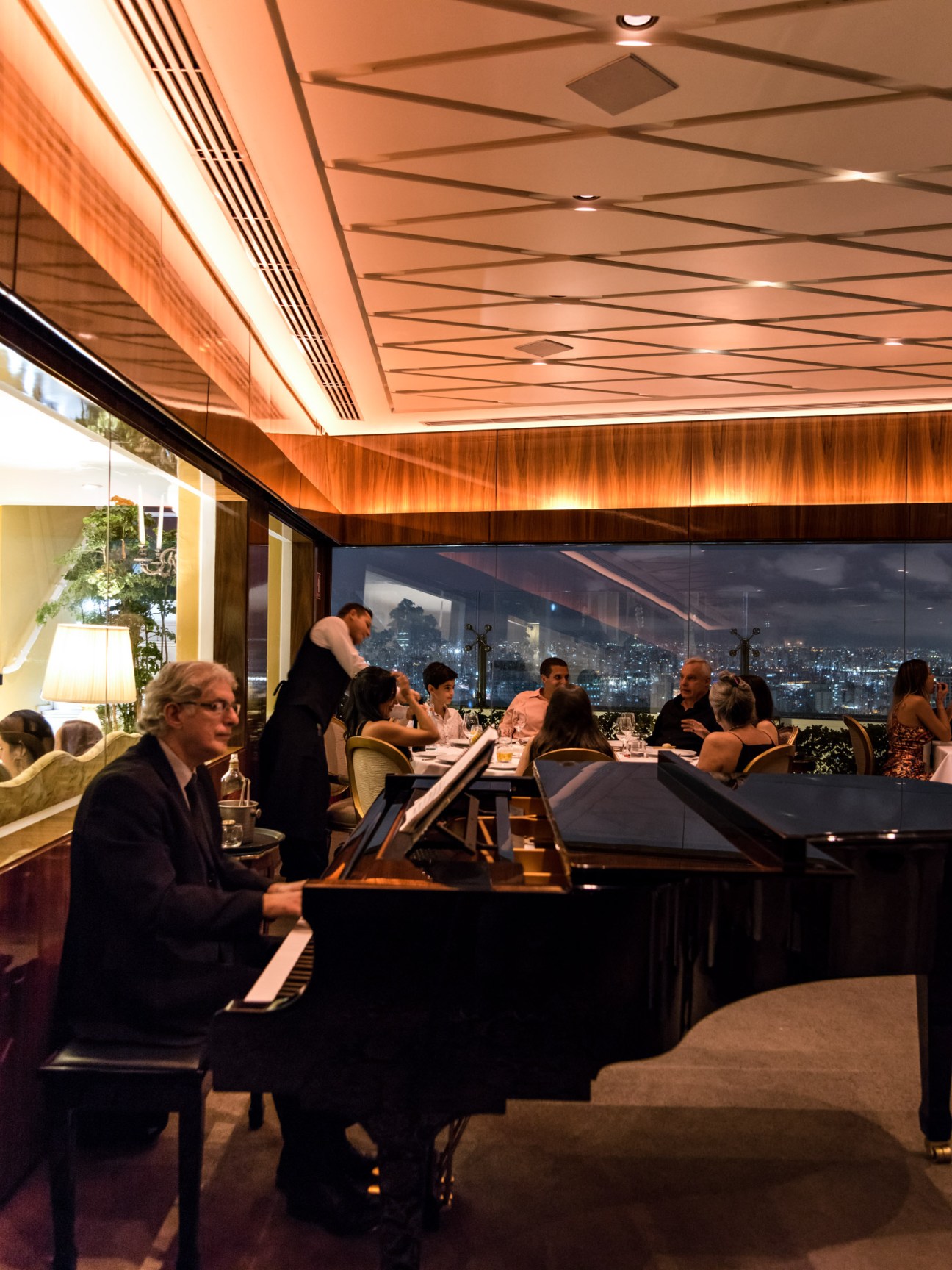 Daniel Casaniga at grand piano in ornate Terraço Itália restaurant with crystal chandeliers and São Paulo skyline view.