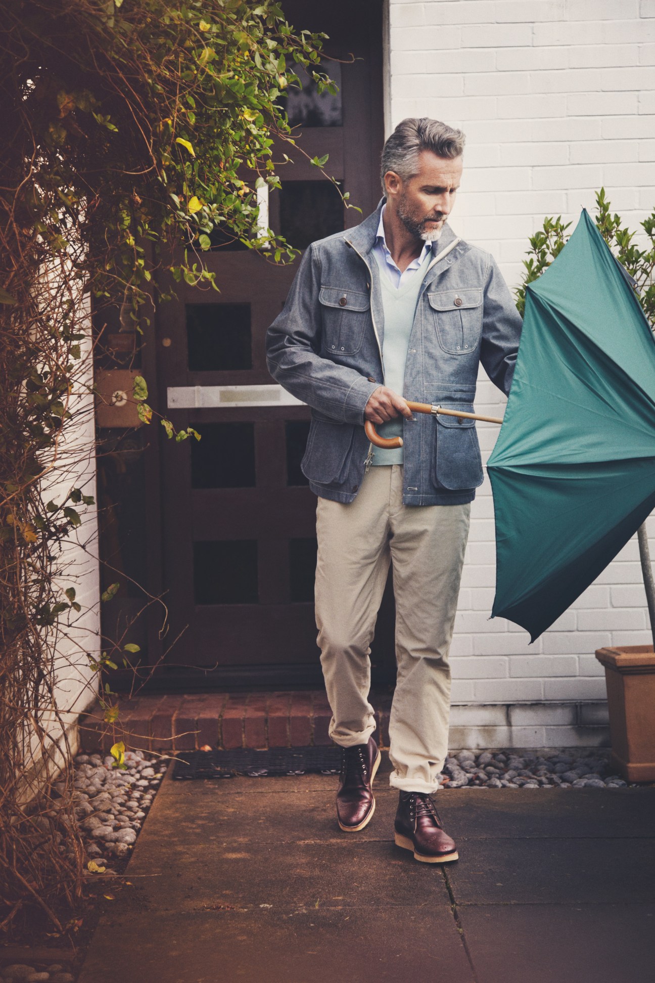 Man in denim jacket and cream trousers holding a teal umbrella outside a modern house.