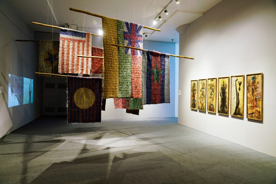 Gallery installation with colorful woven textile artworks suspended from brass rods and framed paintings on white walls.