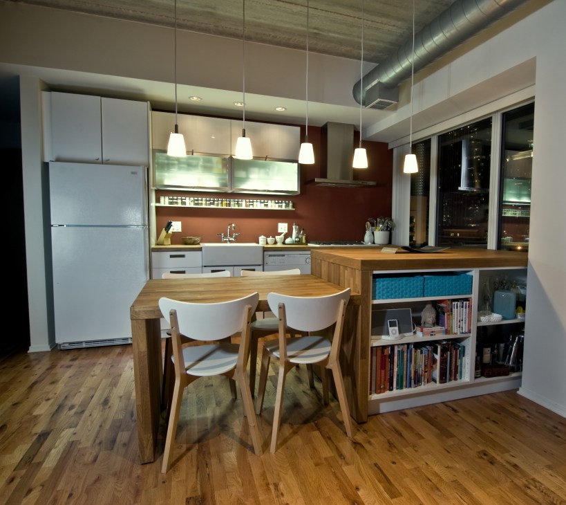 Modern kitchen and dining area with wooden table, white chairs, pendant lights, and open shelving with books.