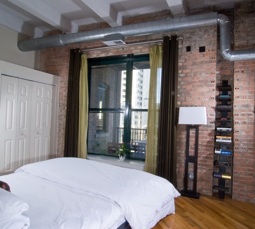 Bedroom with exposed brick walls, white ceiling beams, industrial ductwork, and large windows with yellow curtains.
