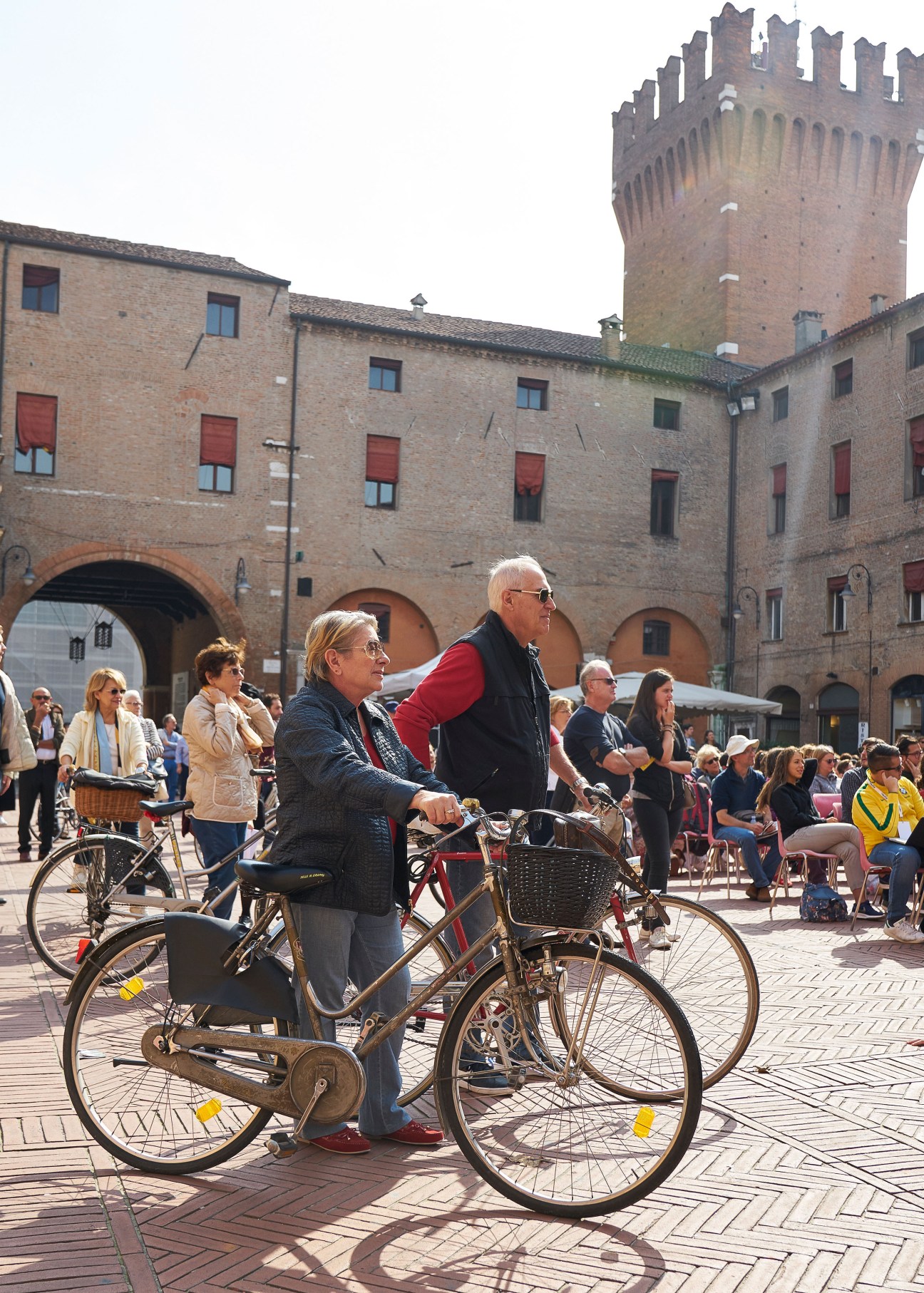 Two cyclists ride through Ferrara's Piazza del Municipio with historic brick buildings and tower behind them.