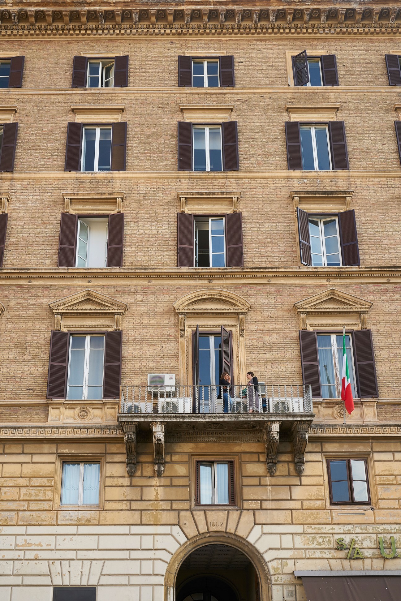 Classical Italian palazzo facade in Rome with tan brick, dark shutters, and Italian flag on balcony.