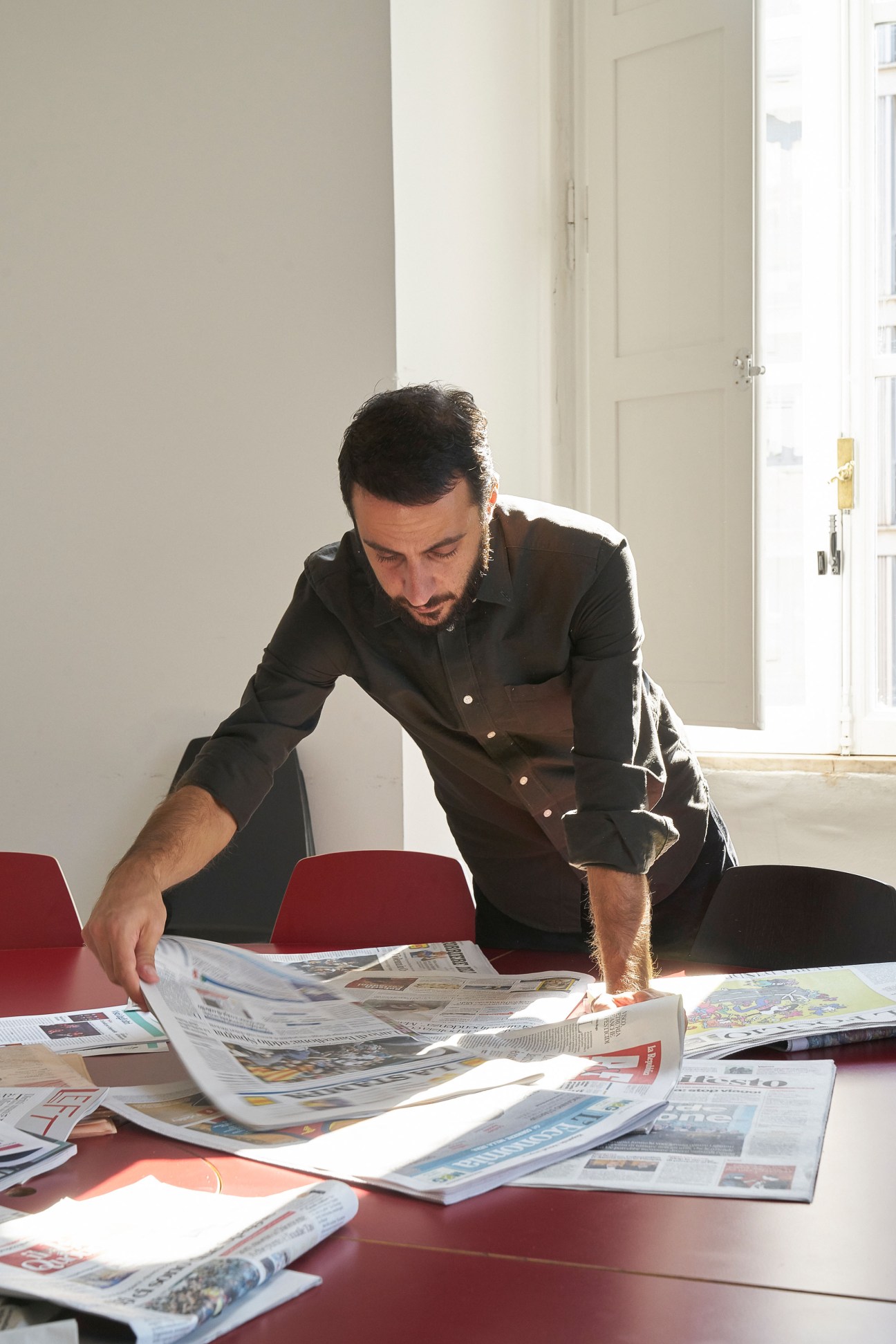 Man in black shirt reviewing international newspapers spread across a red table in bright sunlight.