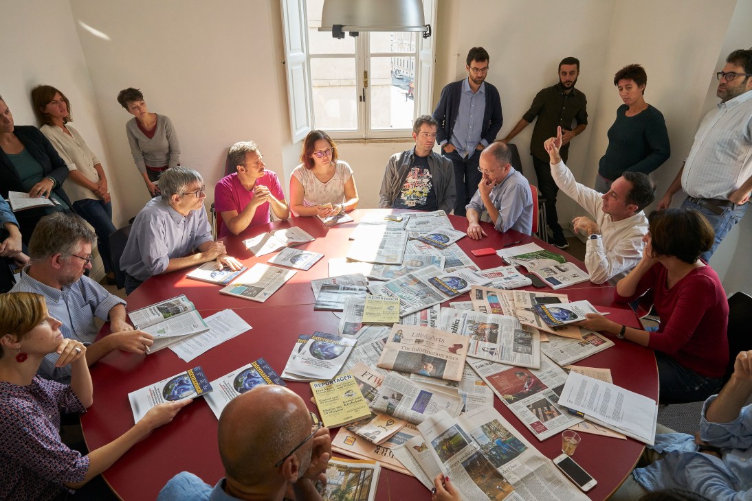 Editorial team gathered around a red table covered with international newspapers and magazines during their Monday morning meeting.