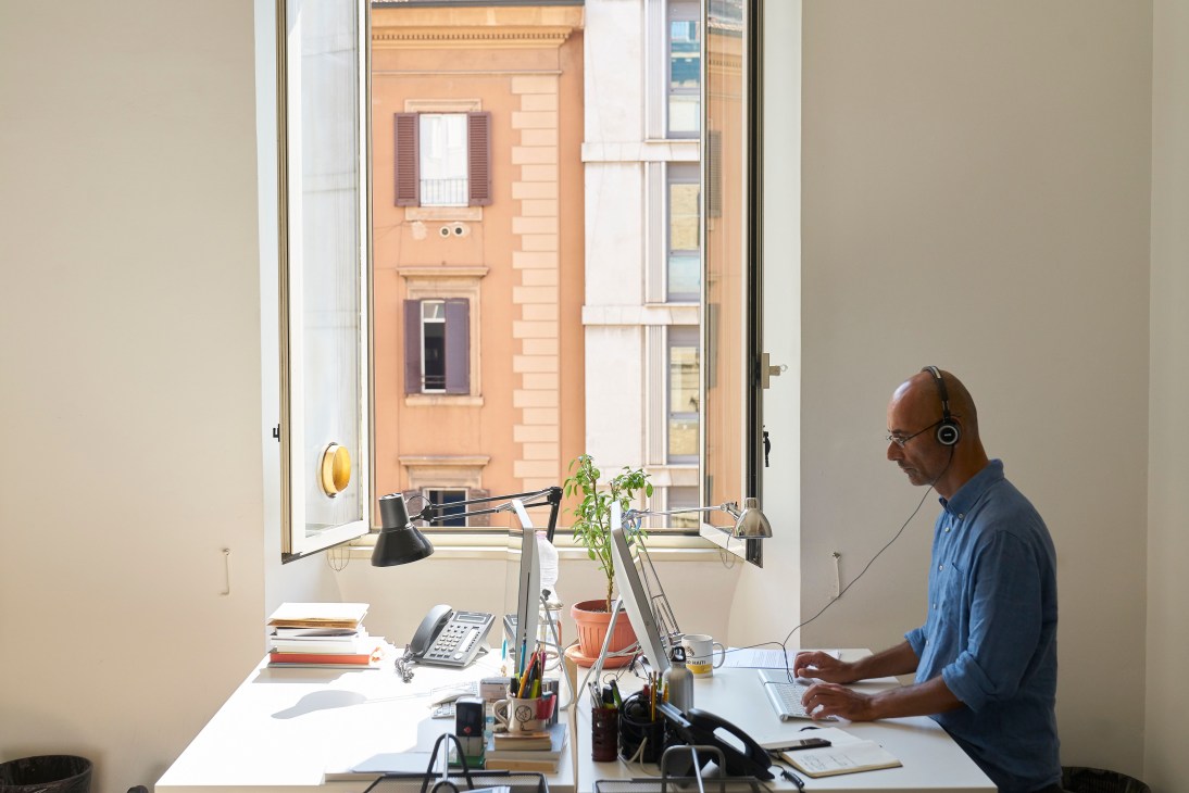 Copy editor Pierfrancesco Romano working at his desk with headphones and computer in a Rome office overlooking terracotta buildings.