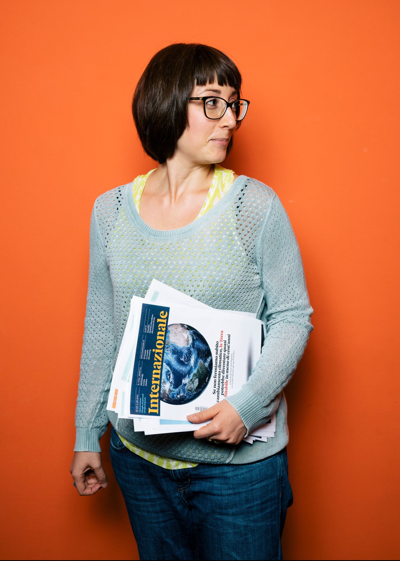 Woman with black bangs and glasses holding Internazionale magazine against orange background.