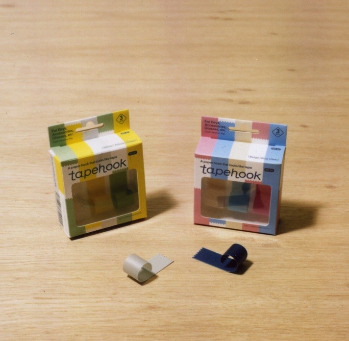 Torafu Architects Tapehook packaging in yellow and multicolored boxes with white and navy hook samples on wooden surface.