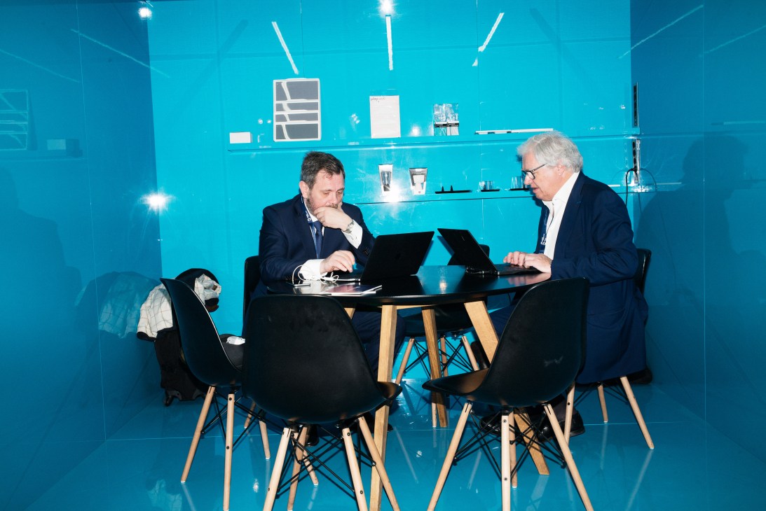 Two businessmen in suits work at laptops at a modern table in a bright turquoise exhibition booth.