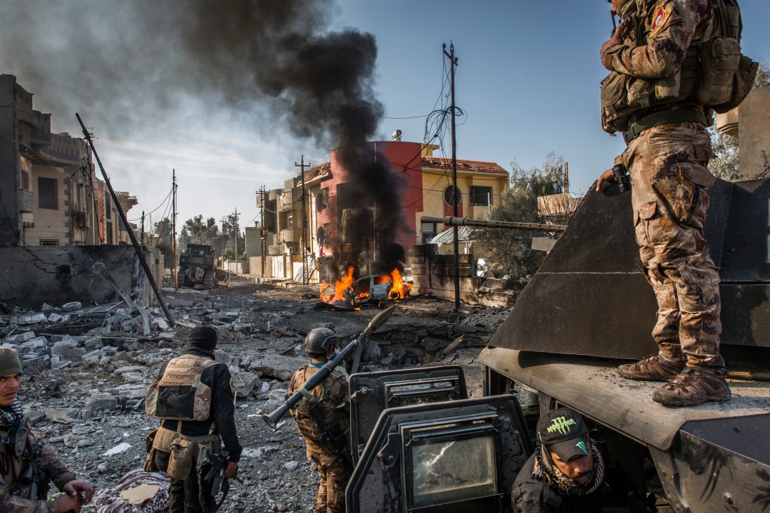 Military personnel observe destruction from an ISIS suicide car-bomb attack in East Mosul, with burning vehicles and damaged buildings.