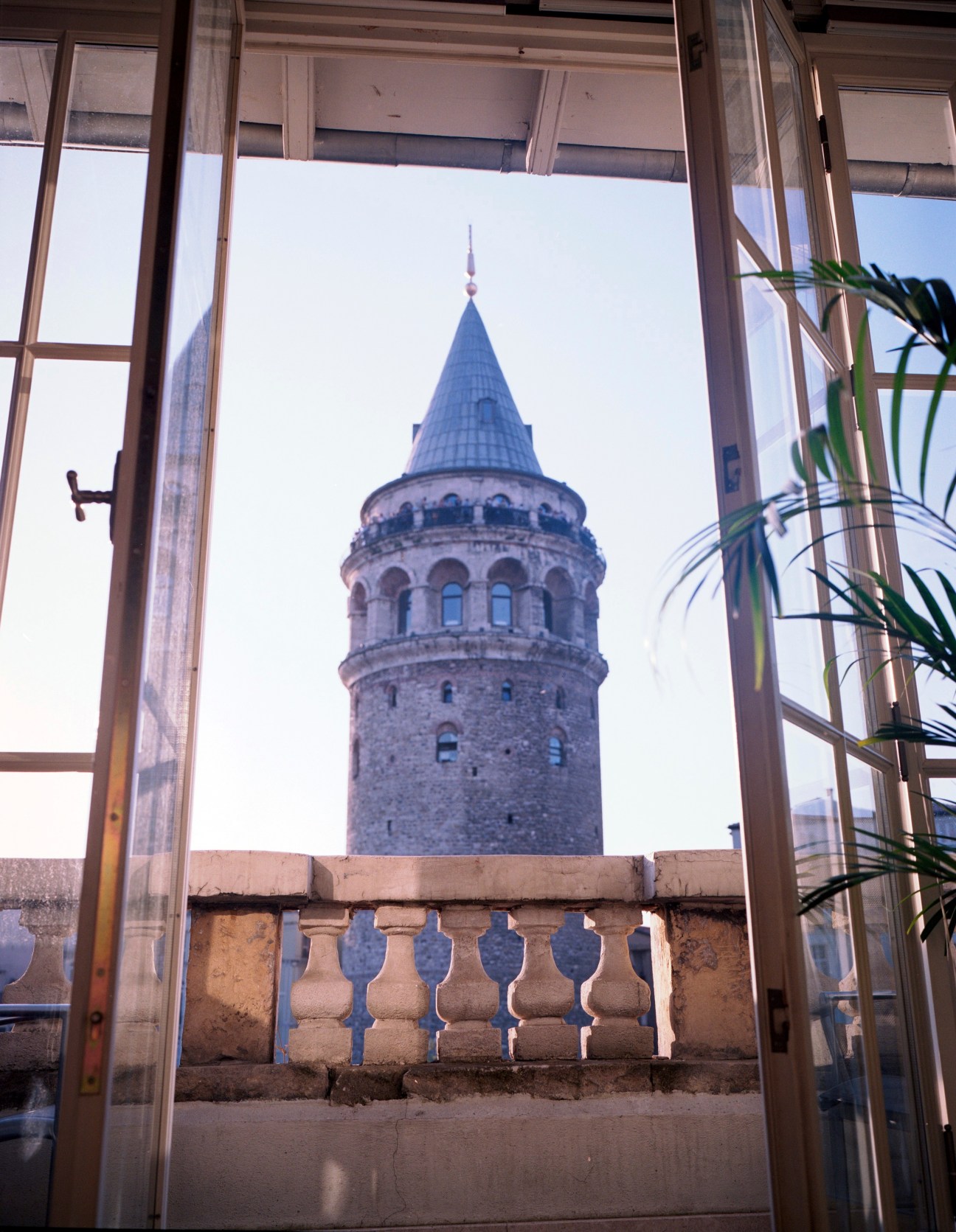View of a historic tower with a conical roof framed through an open window with a potted plant and balustrade visible.