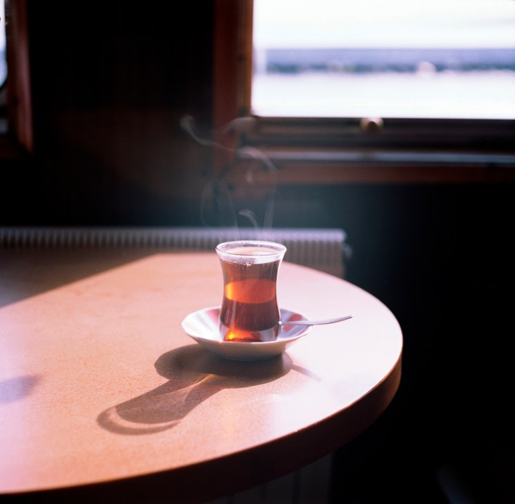 A glass of dark tea on a white saucer with a spoon, placed on a round table with a blurred waterfront view in the background.