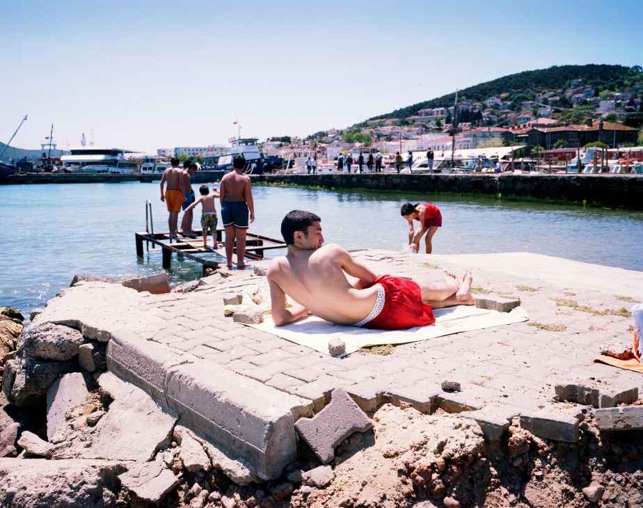 Man lounging on waterfront stone pier with swimmers and boats in Naples harbor background.