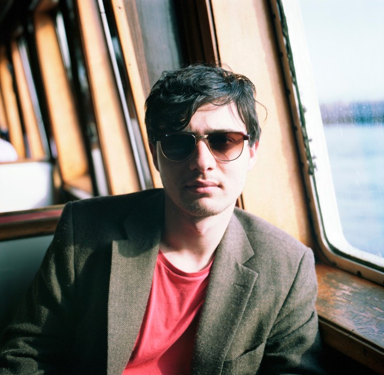 Man wearing sunglasses and a grey blazer on a ferry with water visible through the window behind him.