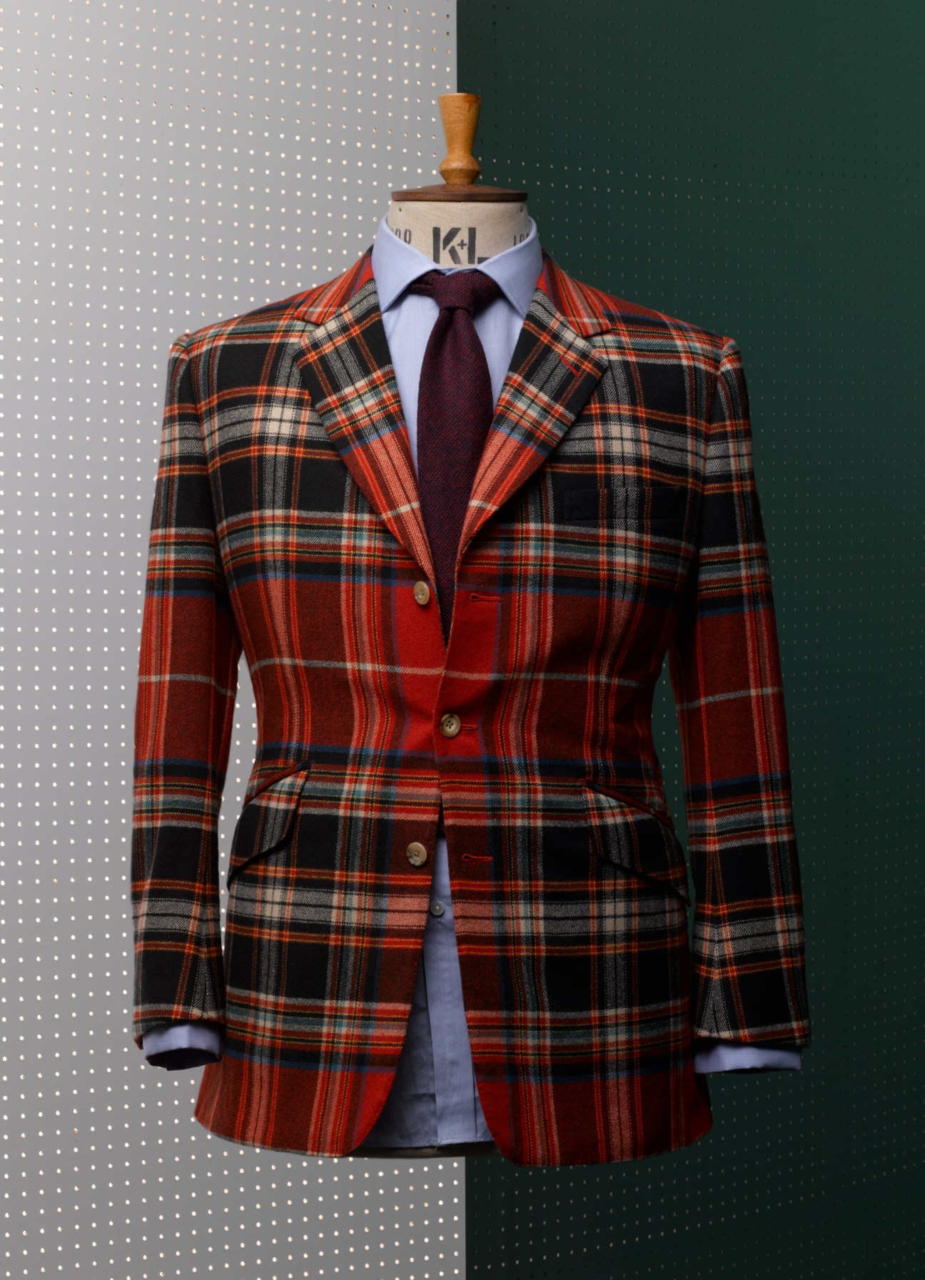 Red and black tartan blazer with burgundy tie displayed on a dress form against a two-tone background.