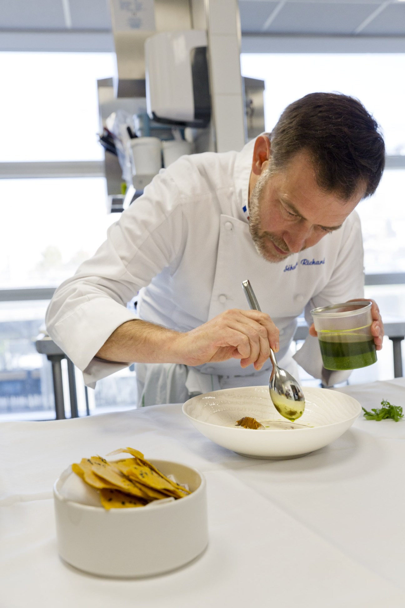 Chef Sébastien Richard in white chef's jacket plating a dish in a bright kitchen.