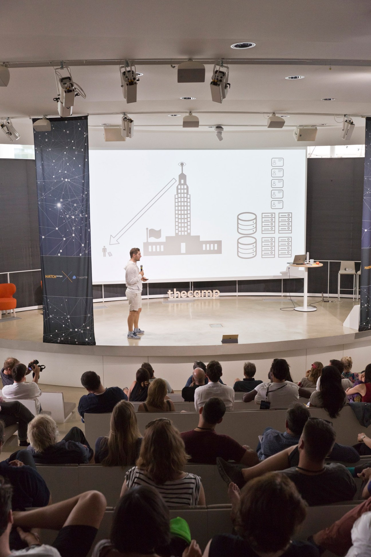 A man speaks at a podium during a Hatch Europe presentation at The Camp, with an audience seated in an auditorium and a large screen displaying urban planning graphics behind him.