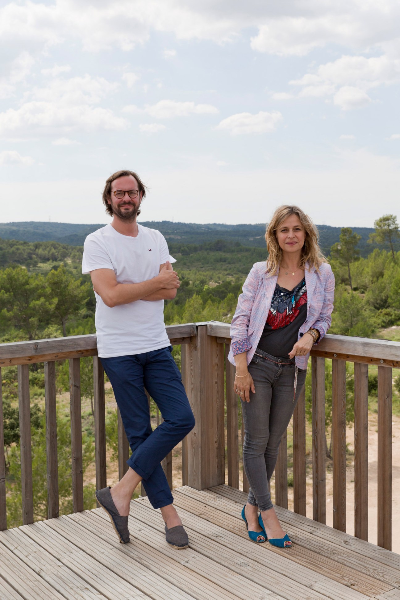 Two people stand on a wooden deck overlooking forested hills under a cloudy sky.
