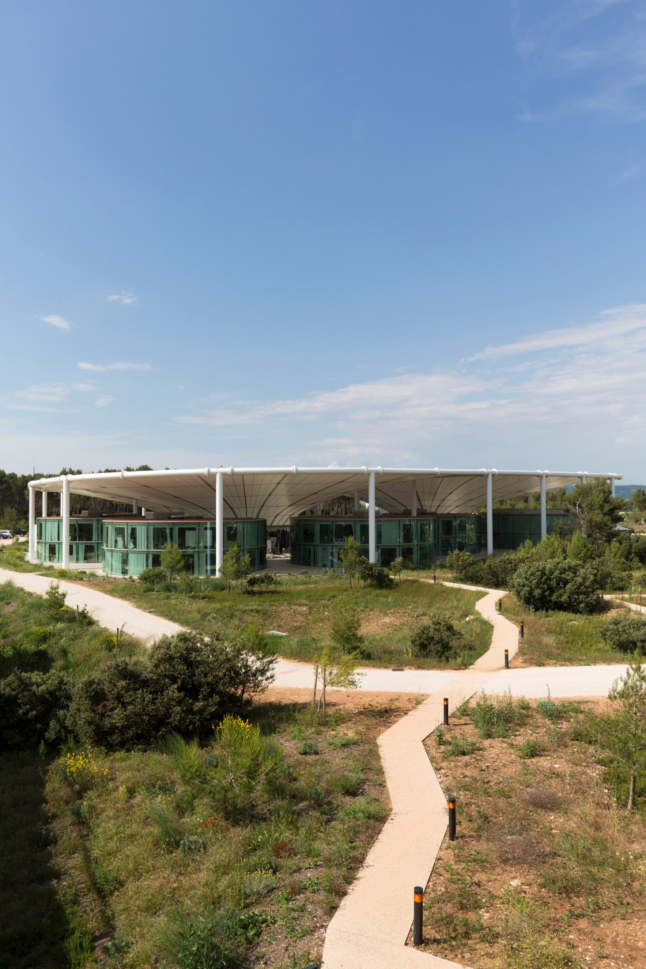 The Camp's modern architecture with white columns and curved metallic canopy sheltering green-tinted glass buildings surrounded by Mediterranean landscape.