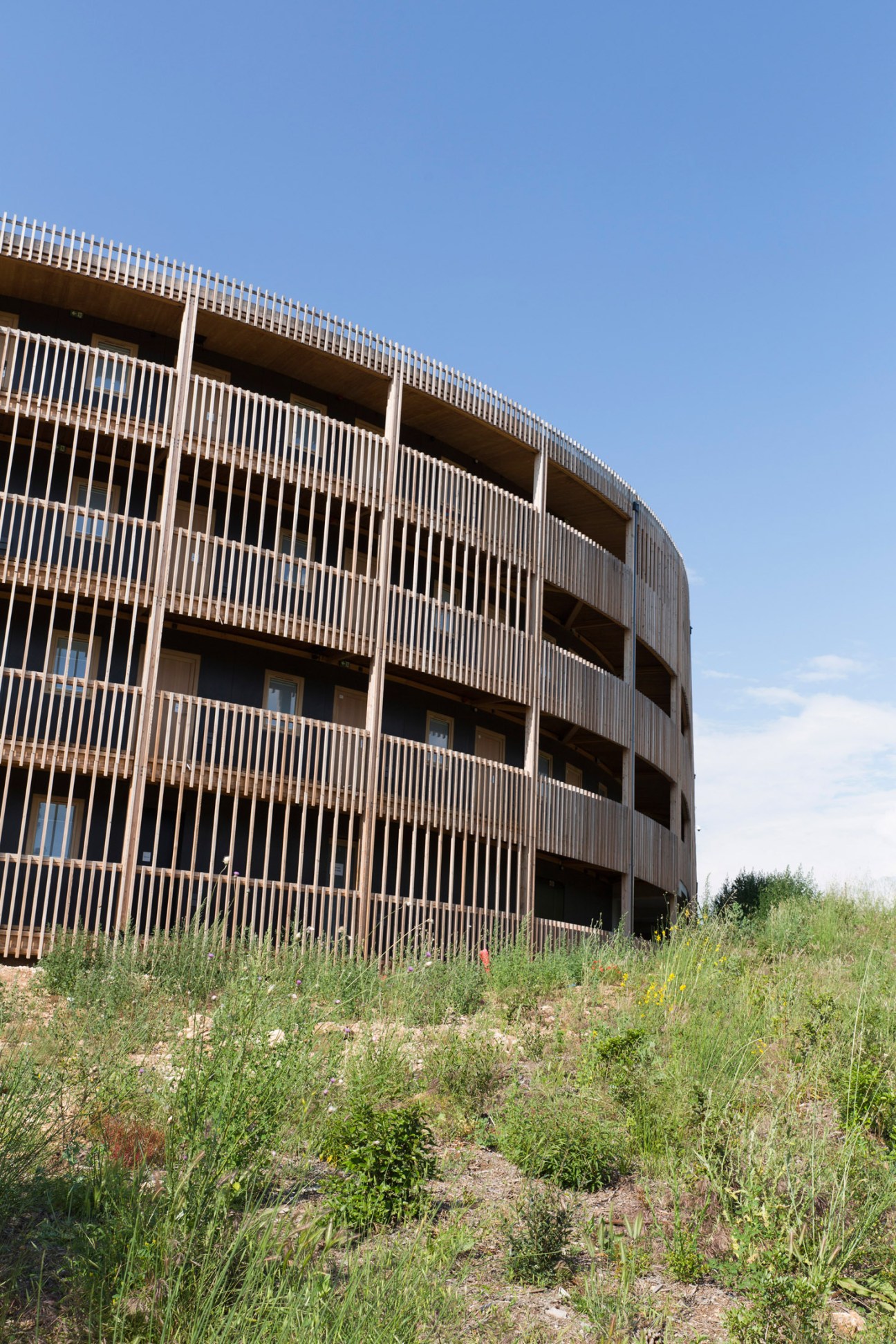 Modern curved building with vertical white slats and multiple levels surrounded by overgrown vegetation.