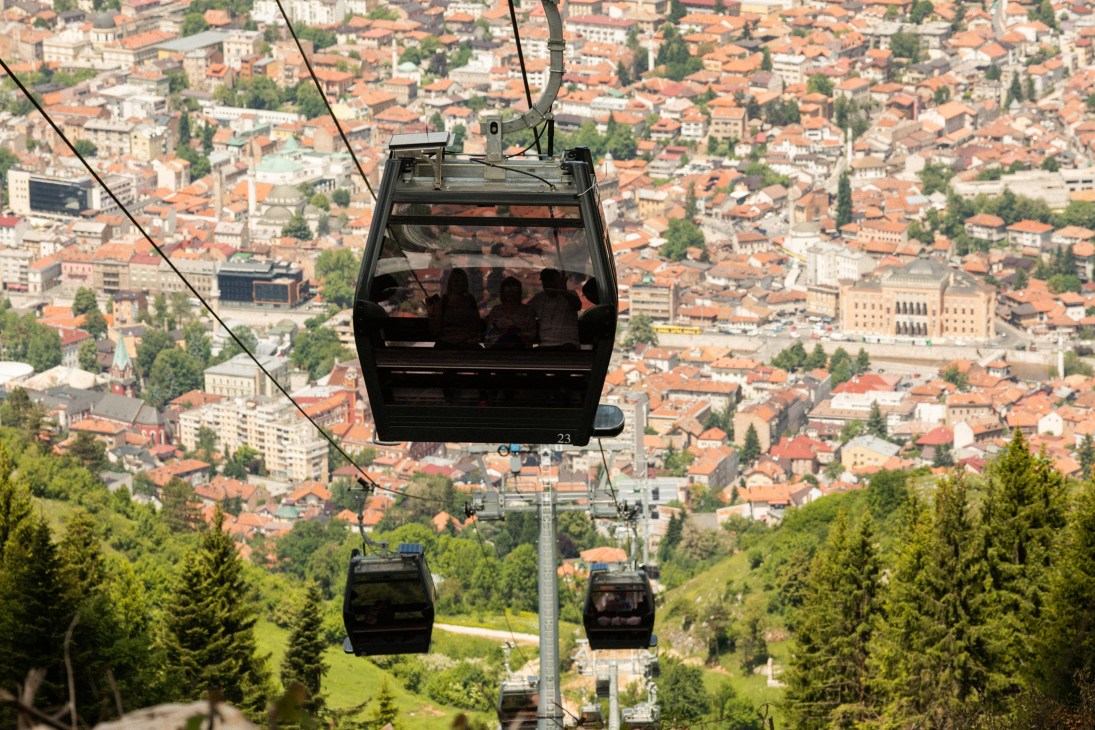 A black cable car gondola suspended above Sarajevo, with the city's red-tiled rooftops and minarets visible below.