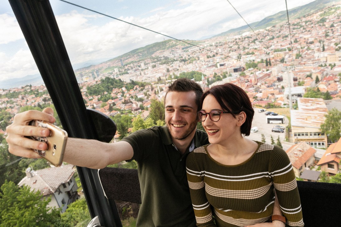 A young couple takes a selfie inside a cable car gondola above Sarajevo's cityscape.