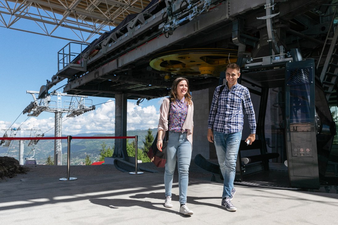 Two young people walk across a mountain-top platform with Sarajevo's cityscape visible below them.