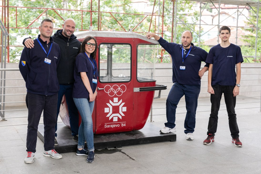 Five staff members posing beside a red Olympic-themed cable car cabin at Sarajevo's station.