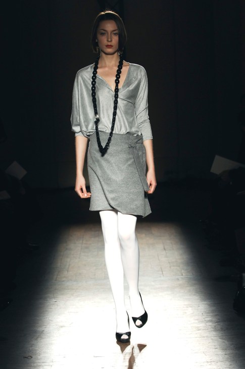 Model wearing Jean-Paul Knott collection with silver-grey blouse, grey skirt, white stockings, black platform heels and black beaded necklace on runway.
