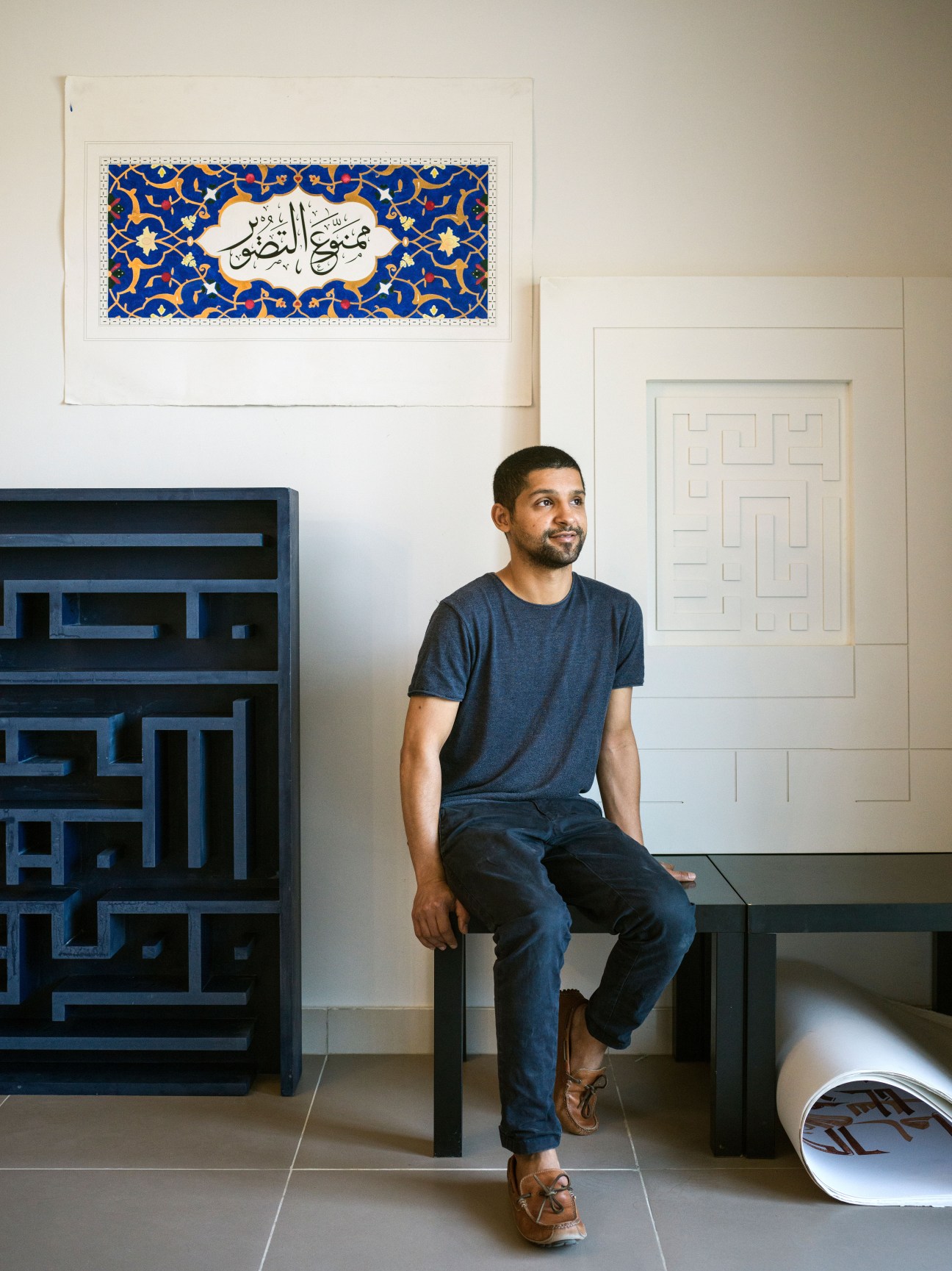 Artist Nasser Al-Salem sits in a studio space surrounded by his artwork including blue geometric sculptures and calligraphic pieces.