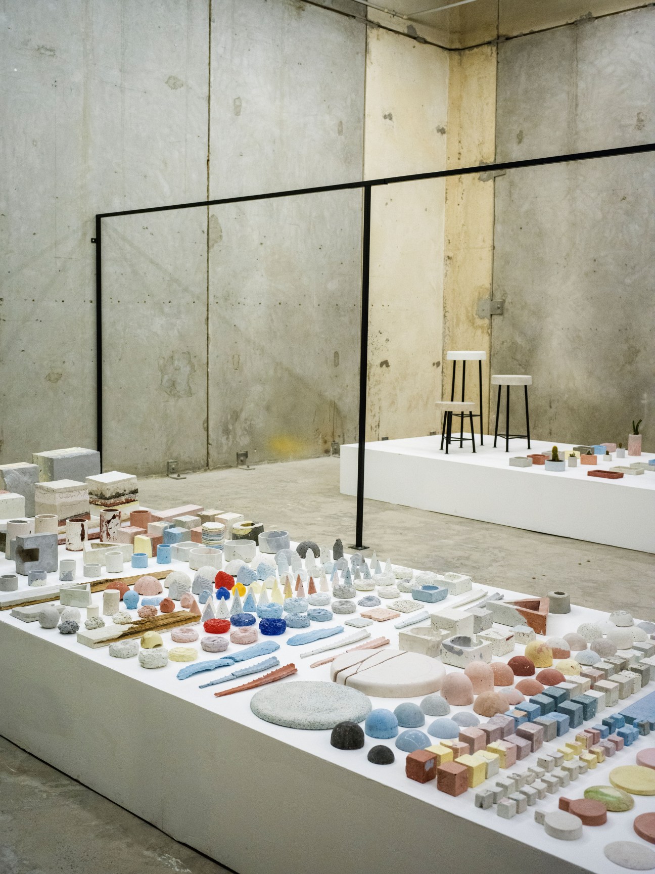 Installation view of colorful ceramic and stone samples displayed on white platforms in a gallery space with industrial concrete walls.