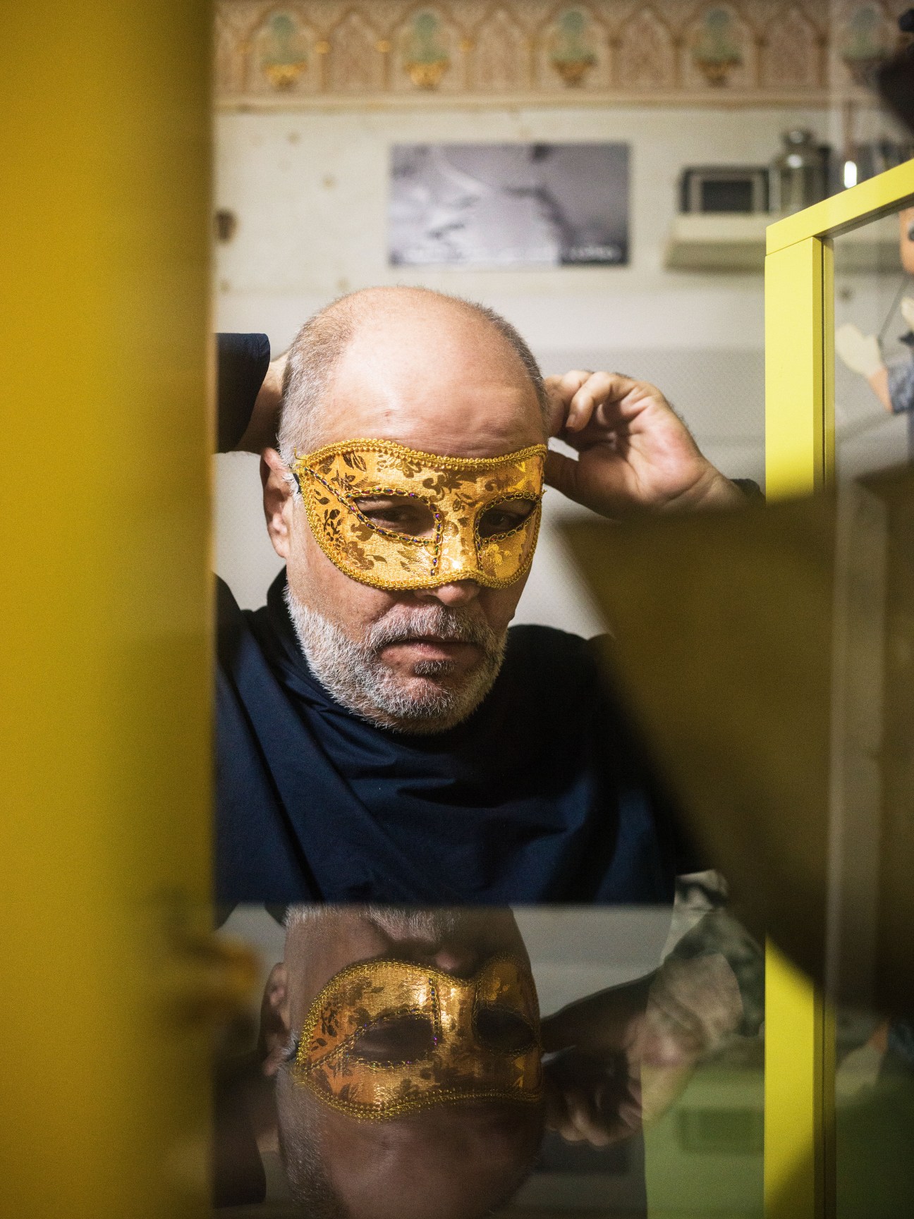 Man wearing a gold venetian mask reflected in a mirror with yellow frame in an art installation.