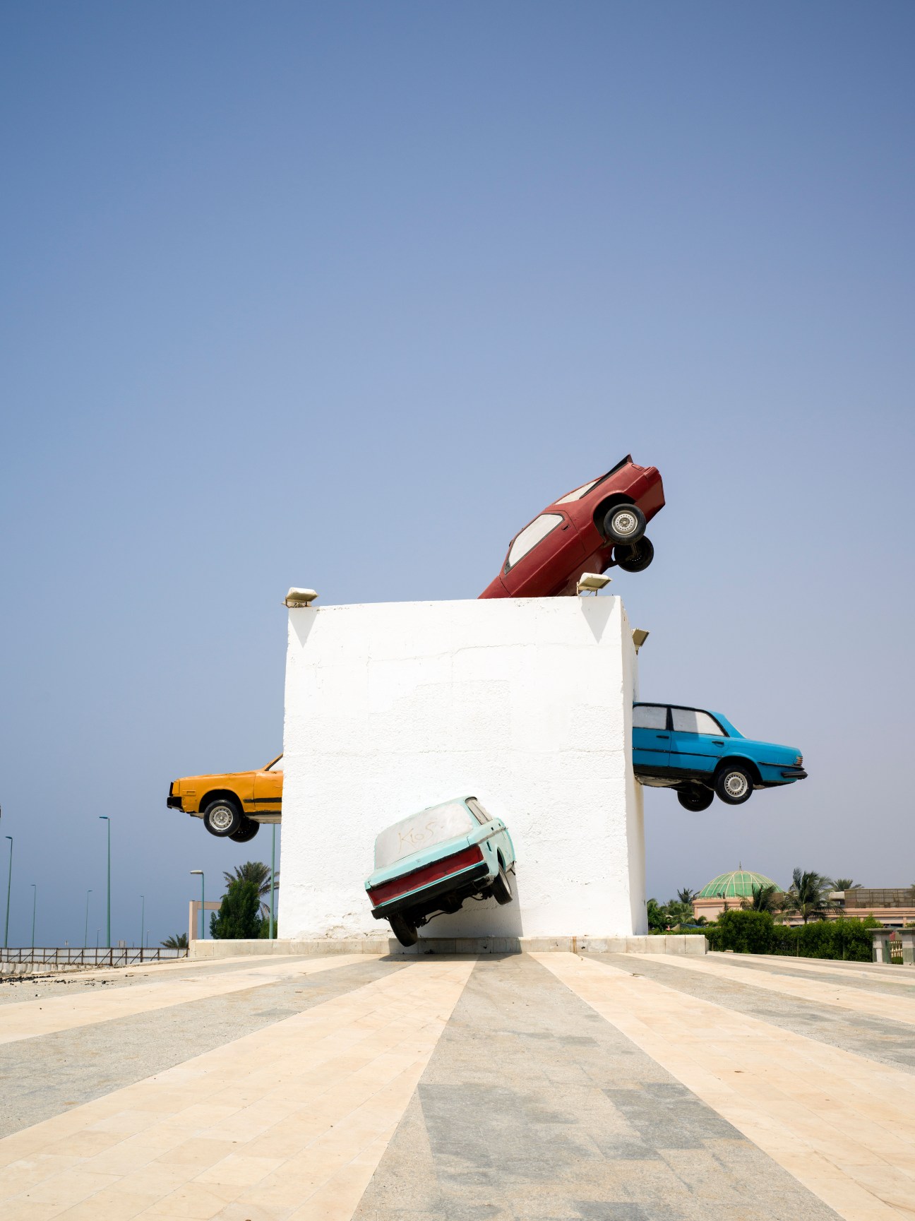 Sculpture of colorful cars appearing to crash through a white rectangular structure against a blue sky.