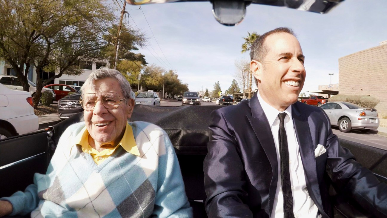 Jerry Seinfeld and an older man smile while sitting in a convertible car on a sunny day.