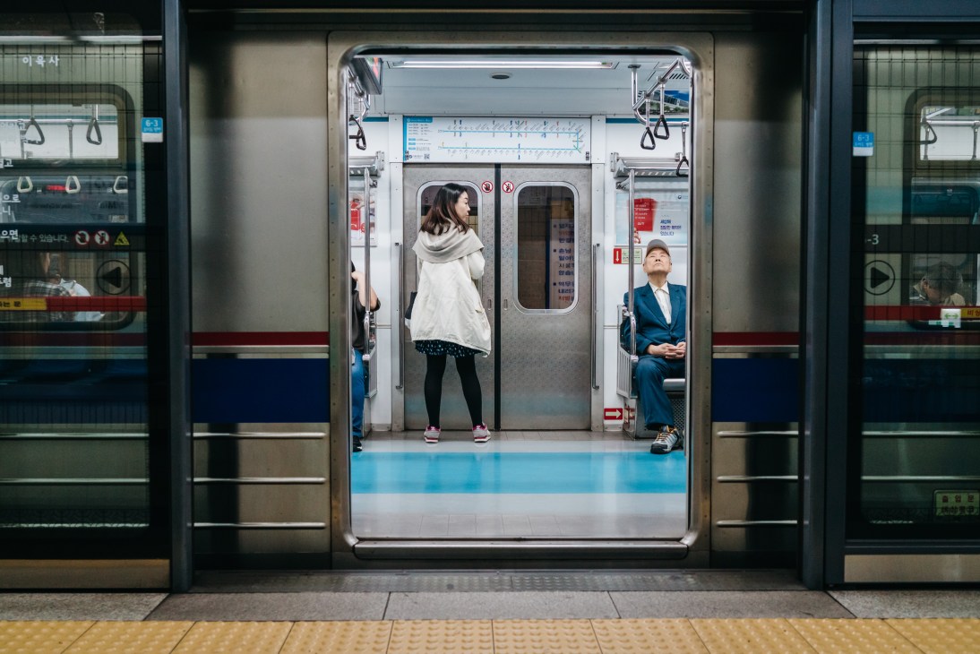 Two passengers inside a clean, modern Seoul subway car with blue and white interior design.