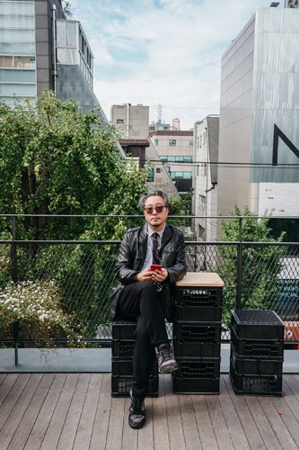 Bernie Cho sits on black crates on a rooftop terrace overlooking a city skyline, wearing sunglasses and a dark jacket.