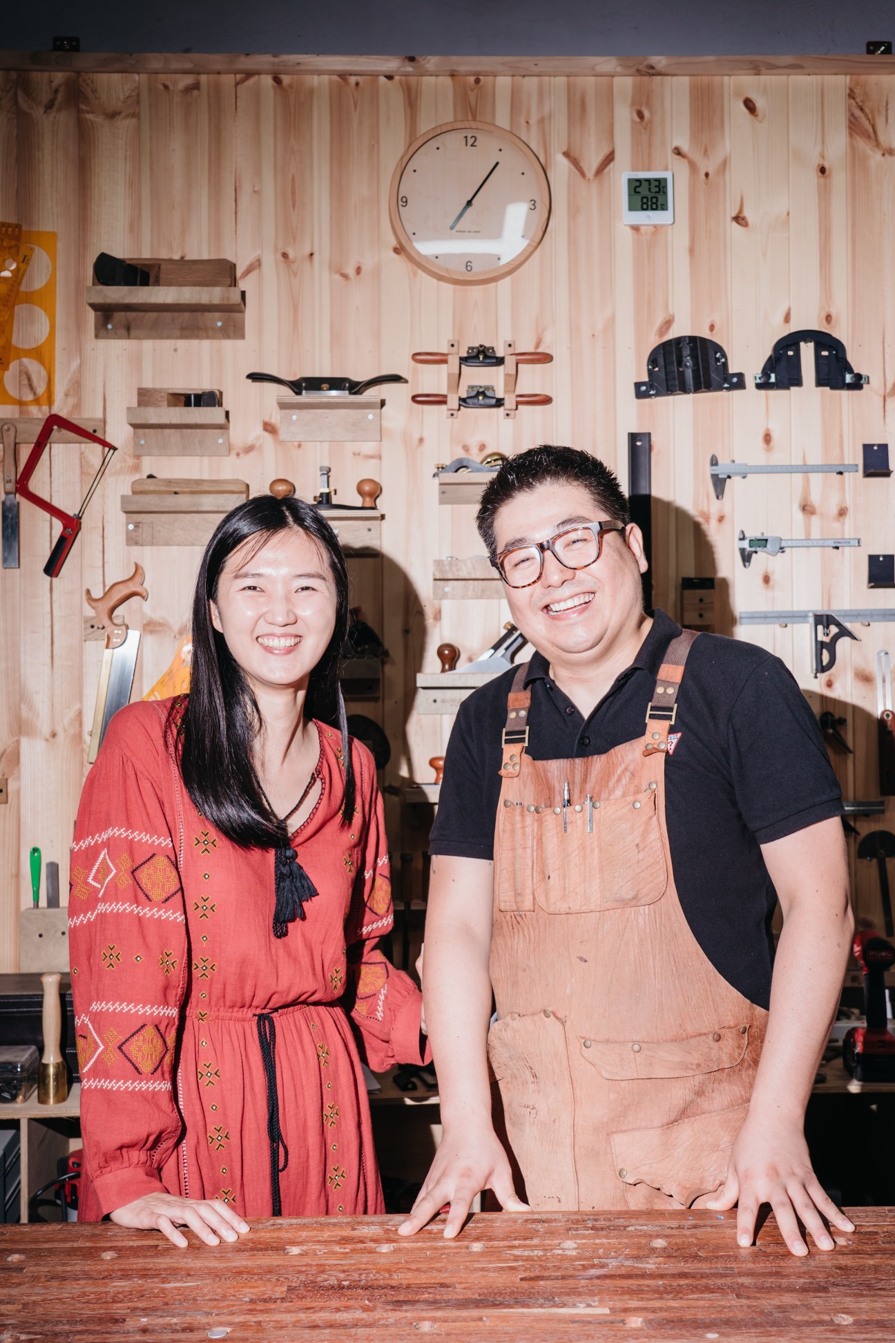 Kim Heeju and Lee Sanggyu standing together in their wooden carpentry workshop with tools on the wall
