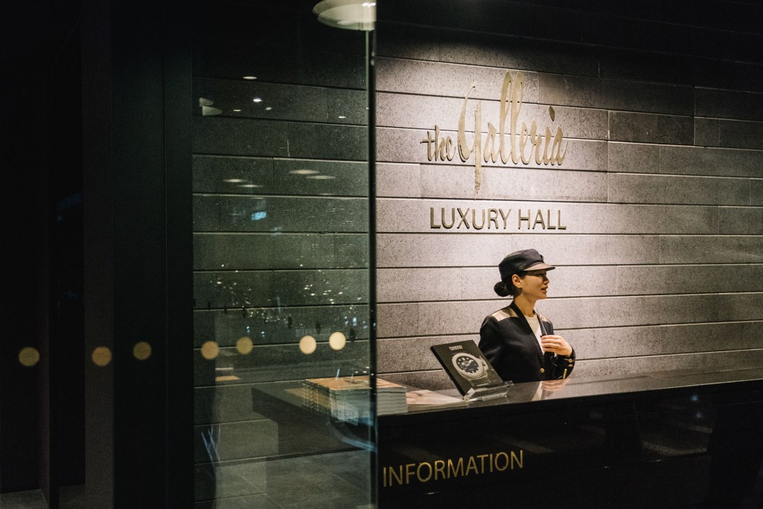 A woman in black uniform and cap stands at an information desk beneath signage reading 'the Galleria' and 'LUXURY HALL'.