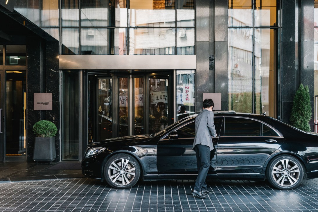 A valet in grey uniform stands beside a black Mercedes sedan outside a modern glass building in Seoul.