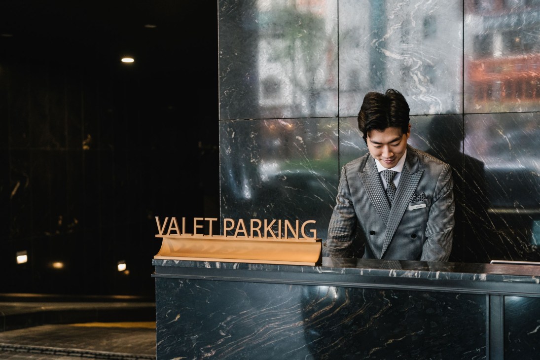 A valet attendant in a grey uniform stands at a reception desk with a 'VALET PARKING' sign.