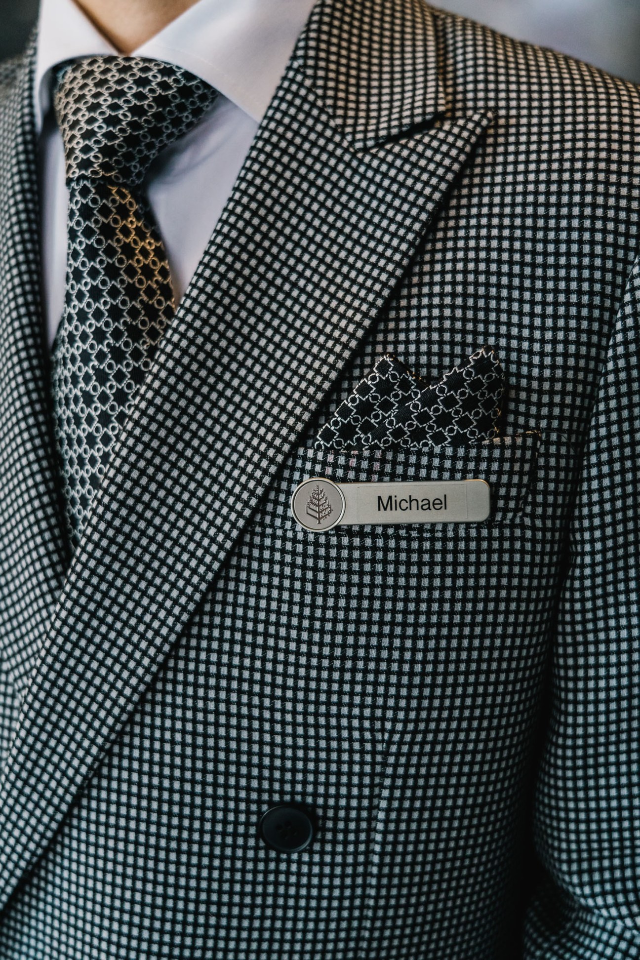 Close-up of a valet's name badge reading 'Michael' pinned to a black and white checkered blazer with patterned tie.